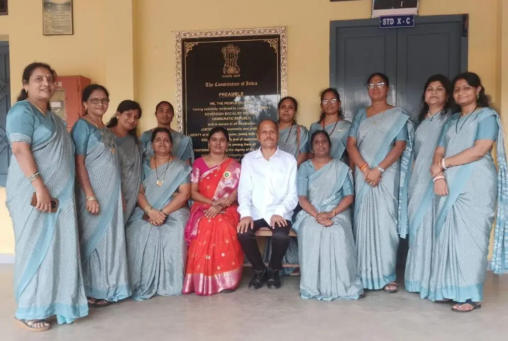 Ms. Devika Alwar, dressed in a bright orange and red saree, seated alongside the Academic Head, Social Development team members, and teaching staff wearing matching light blue sarees in front of a Preamble of the Constitution board at St. Ann's Convent School.